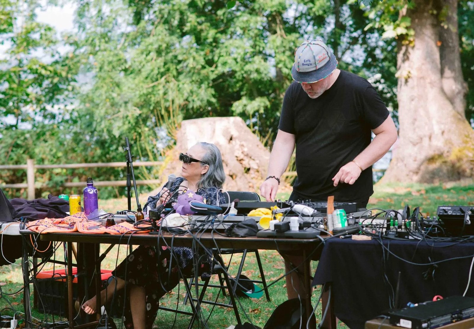 Two artists work with microphones, mixers, and electronic equipment on an outdoor table beneath trees.