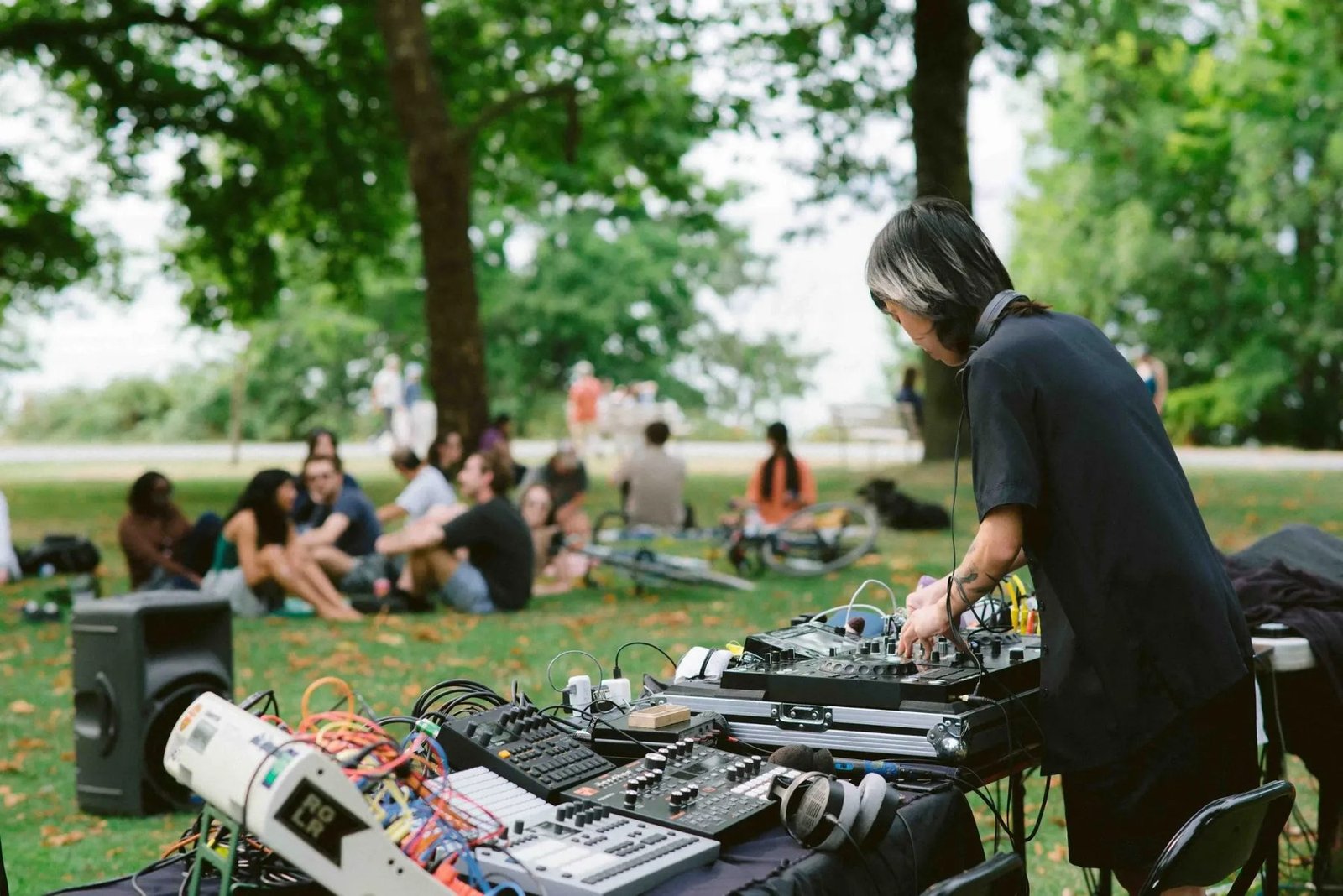 An electronic music performer works at a table of mixers and devices while people gather in the park.
