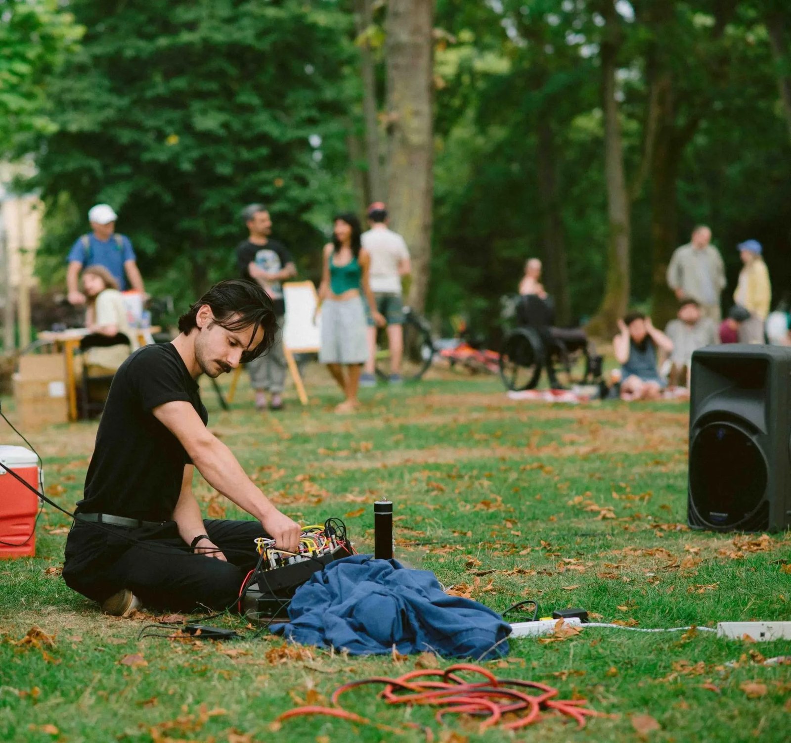 A performer sits on the grass operating a compact electronic instrument setup with the public gathering behind.