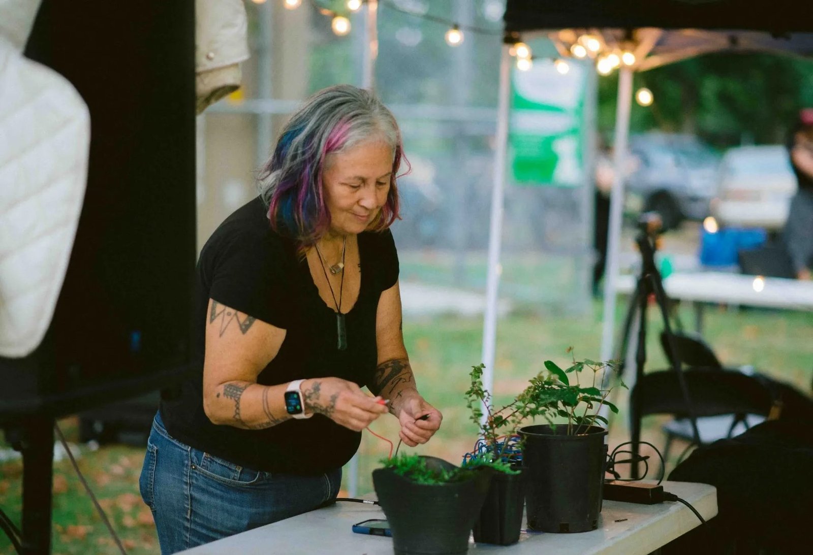 A participant works with small electronic components beside potted plants under an event canopy.