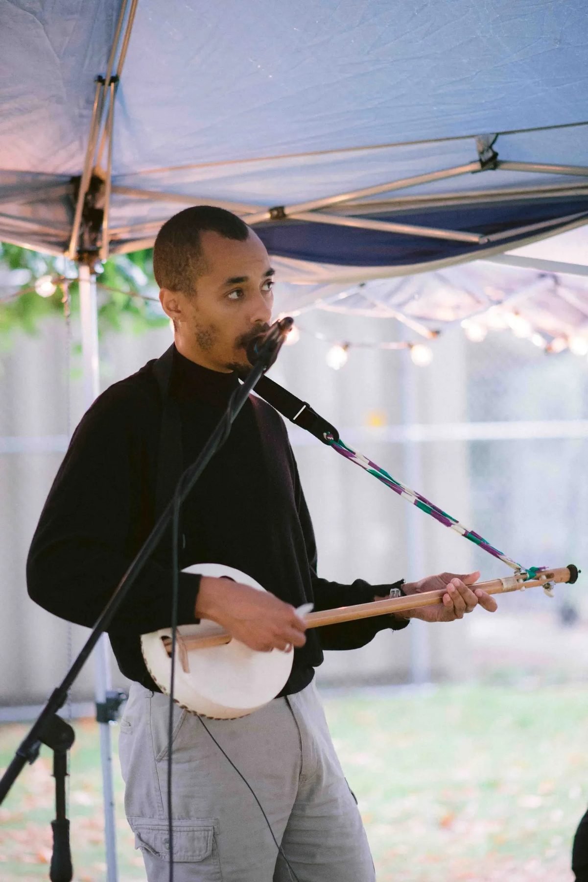 A musician performs into a microphone while playing a long-necked string instrument under an outdoor canopy.