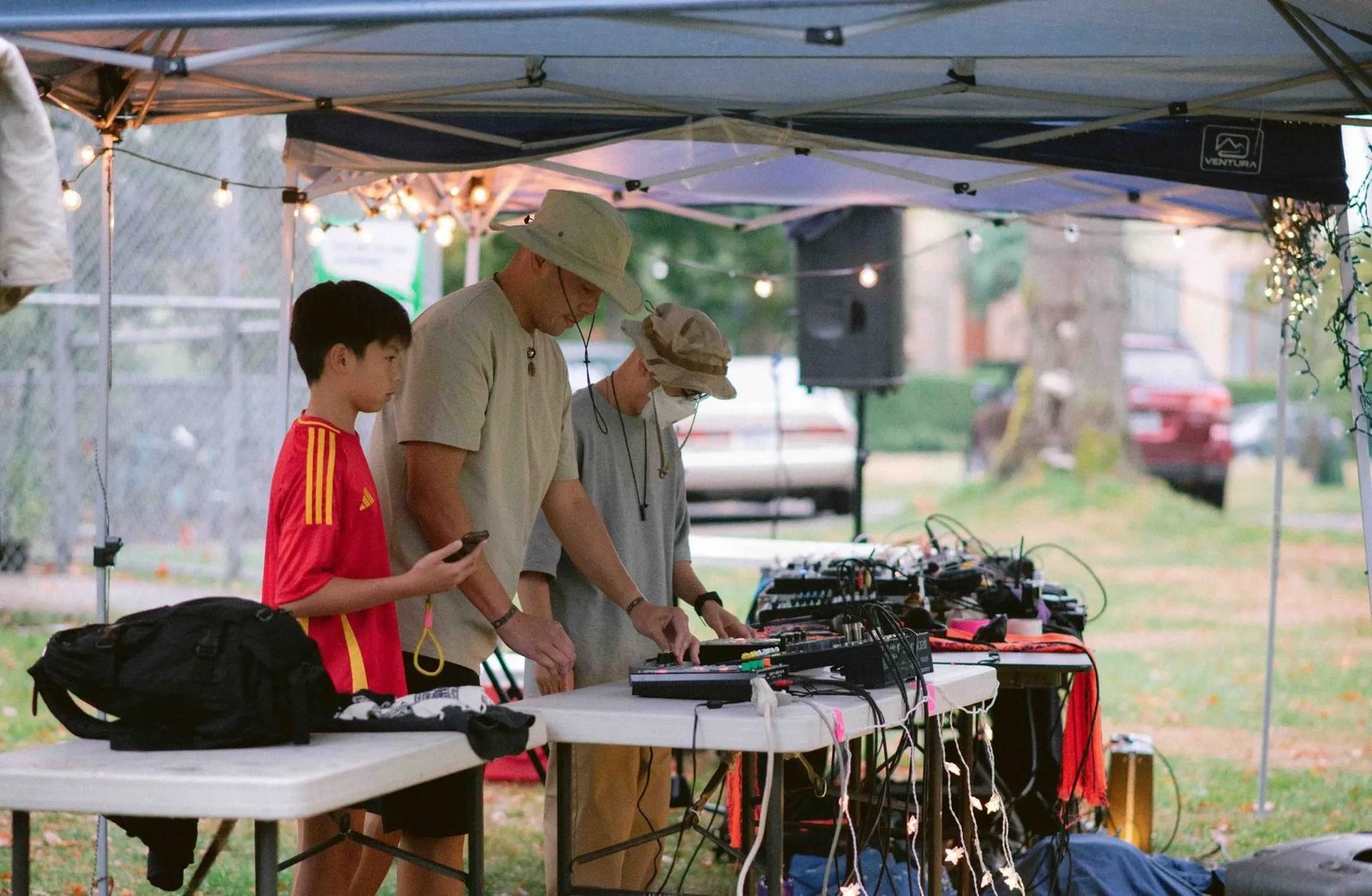 Three participants stand around electronic music equipment on tables beneath an outdoor canopy.