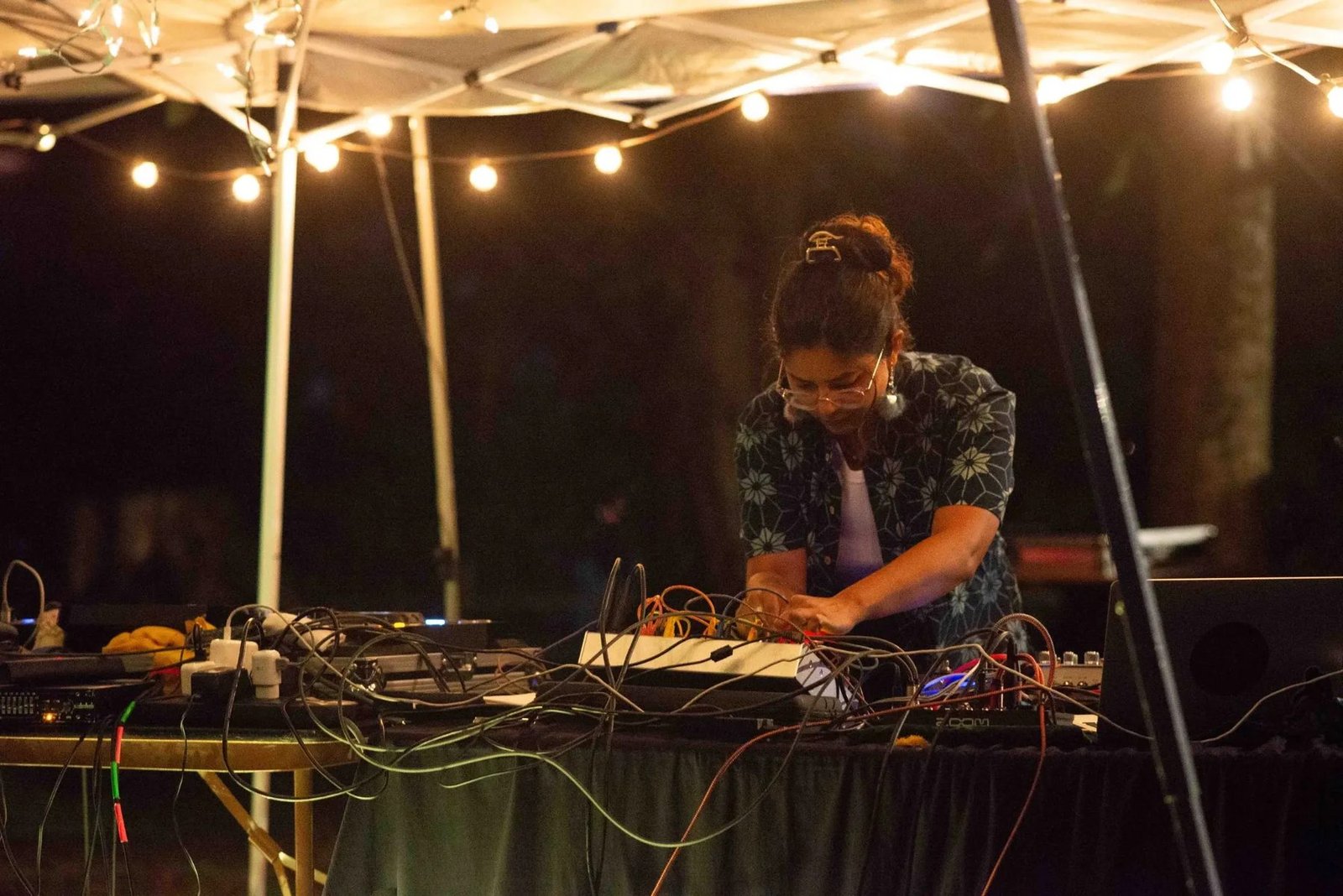 An electronic performer works at a densely cabled equipment table under warm string lights at night.