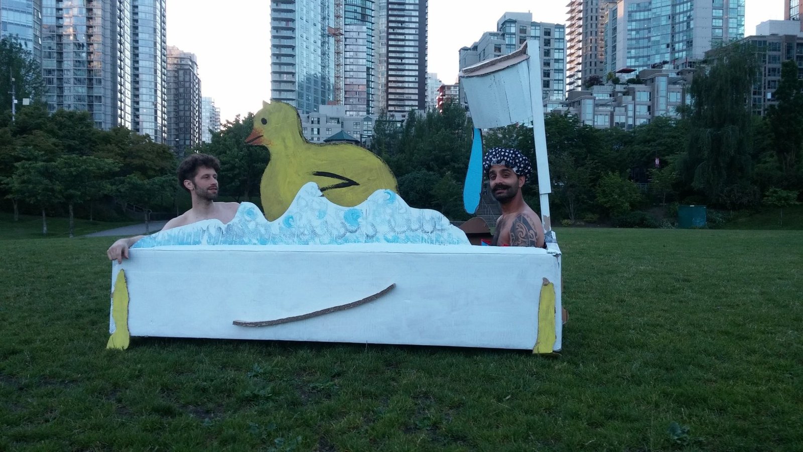 Two participants inside a playful cardboard bath-tub sculpture with a large yellow duck, staged in a grassy Vancouver park with high-rise buildings behind them.