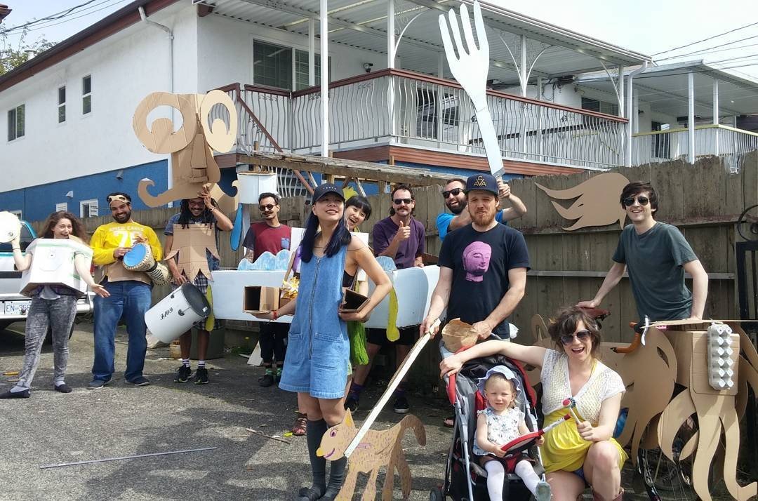 A group of participants pose together with cardboard costumes, parade props, and musical instruments during Cardboard Day.