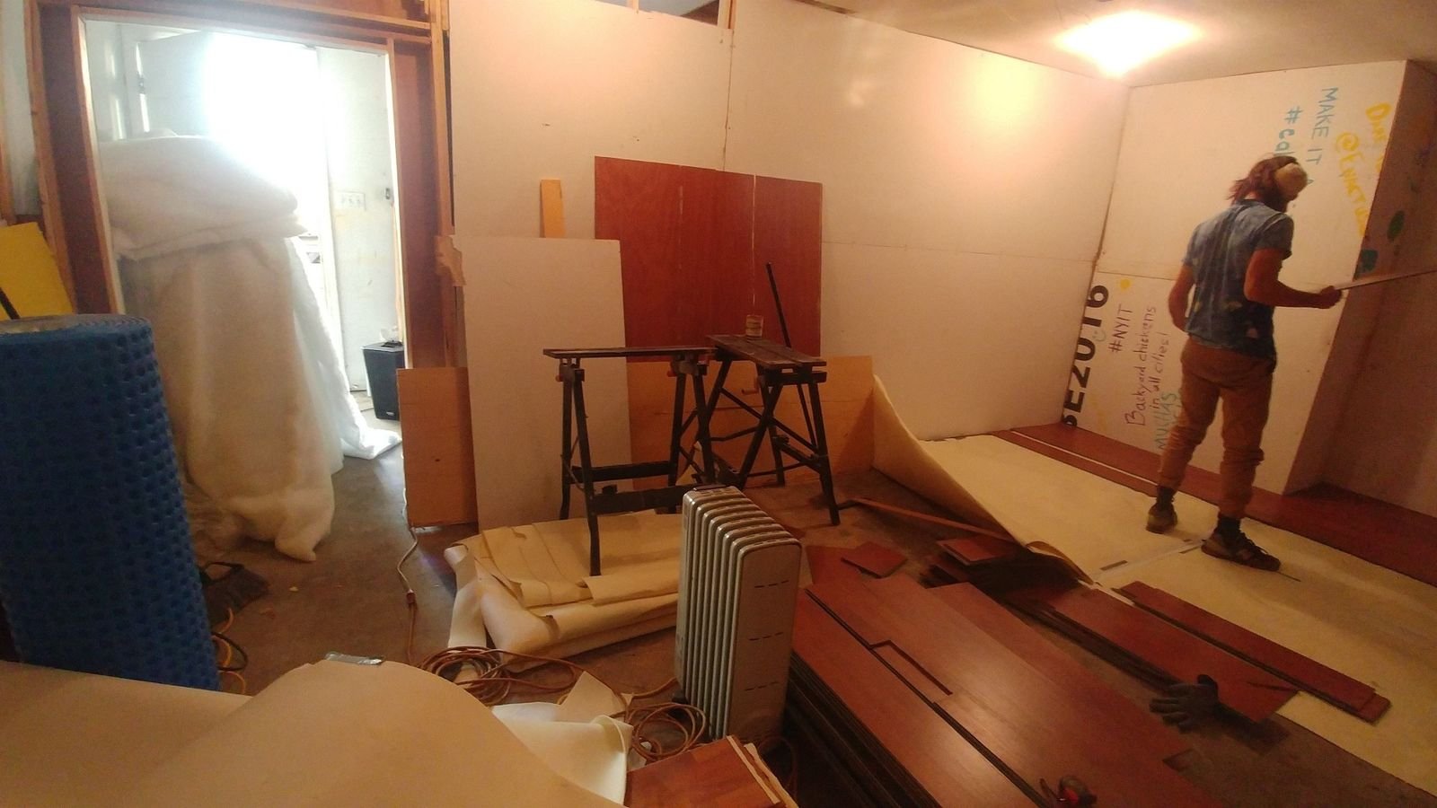 A person works inside the garage among reclaimed flooring, wall panels, insulation, tools, and building materials.