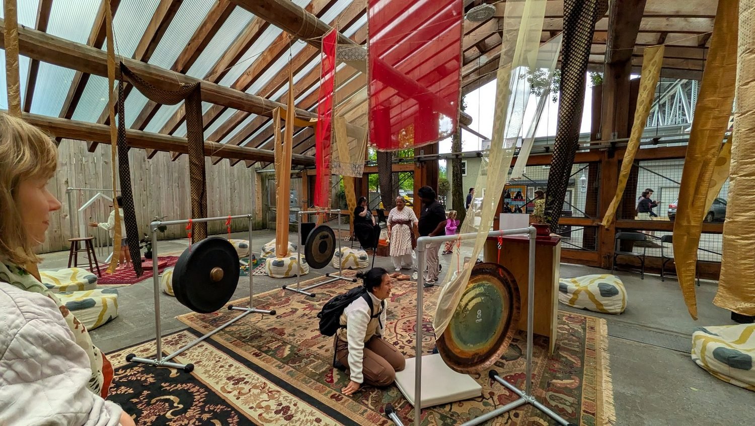 A visitor kneels to play a gong while others sit and stand nearby beneath hanging fabric banners.