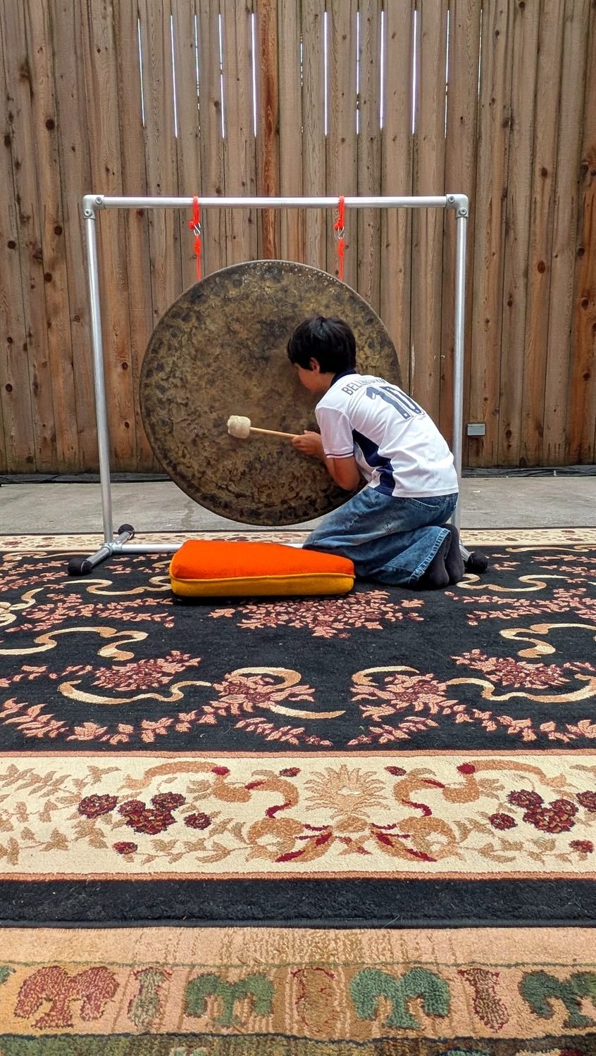 A child kneels close to a large gong, mallet in hand, with an orange cushion beside them.