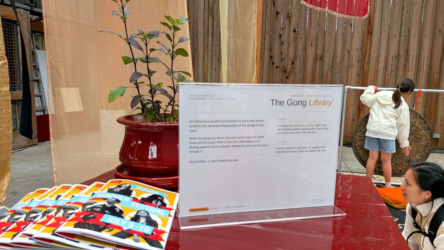 A printed Gong Library information placard and a stack of Indian Summer Festival programs on a table, with a visitor playing a gong in the background.