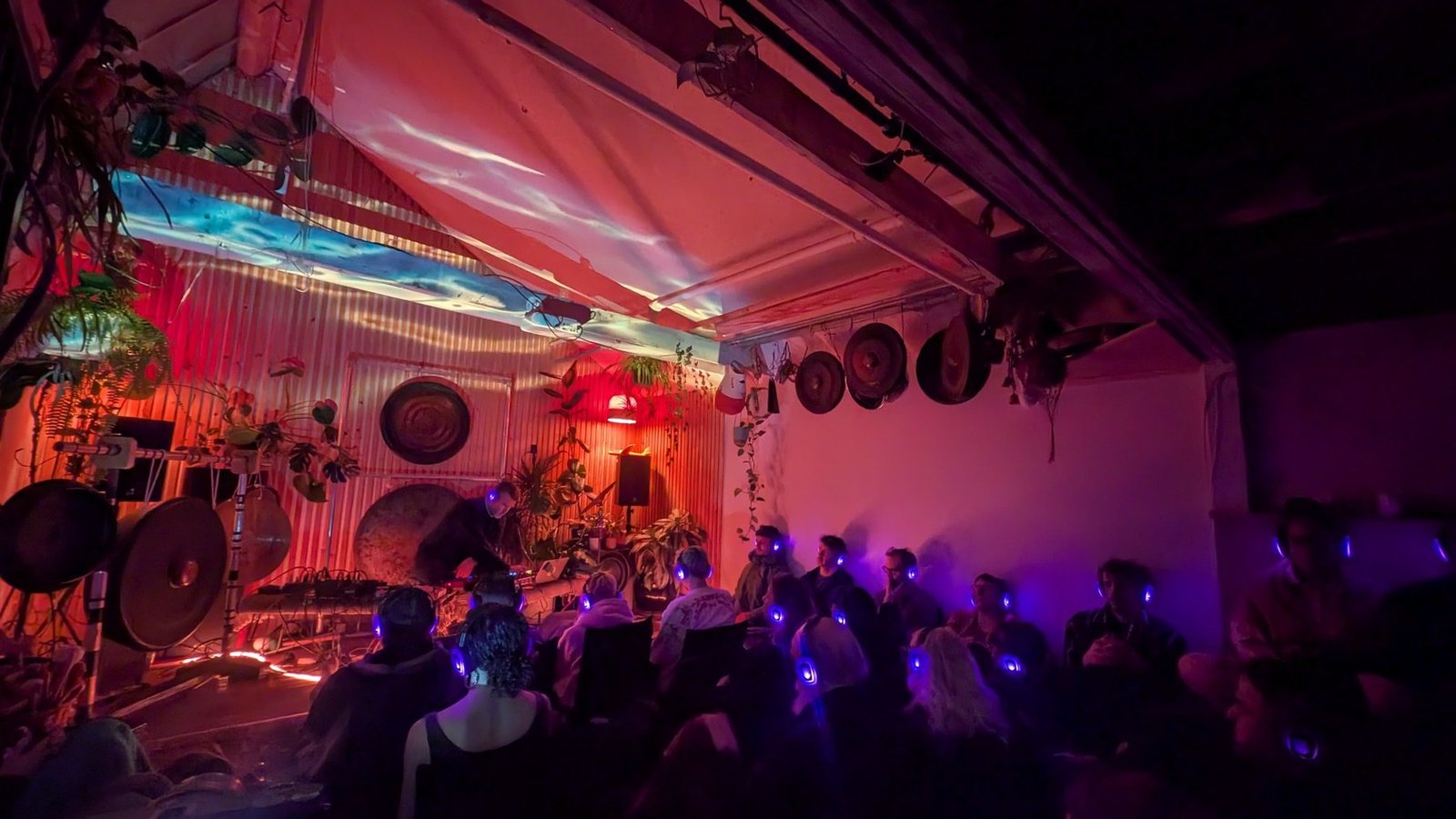 A seated audience wearing illuminated wireless headphones listens to Michael Red performing in the Gong Library under red and purple light.