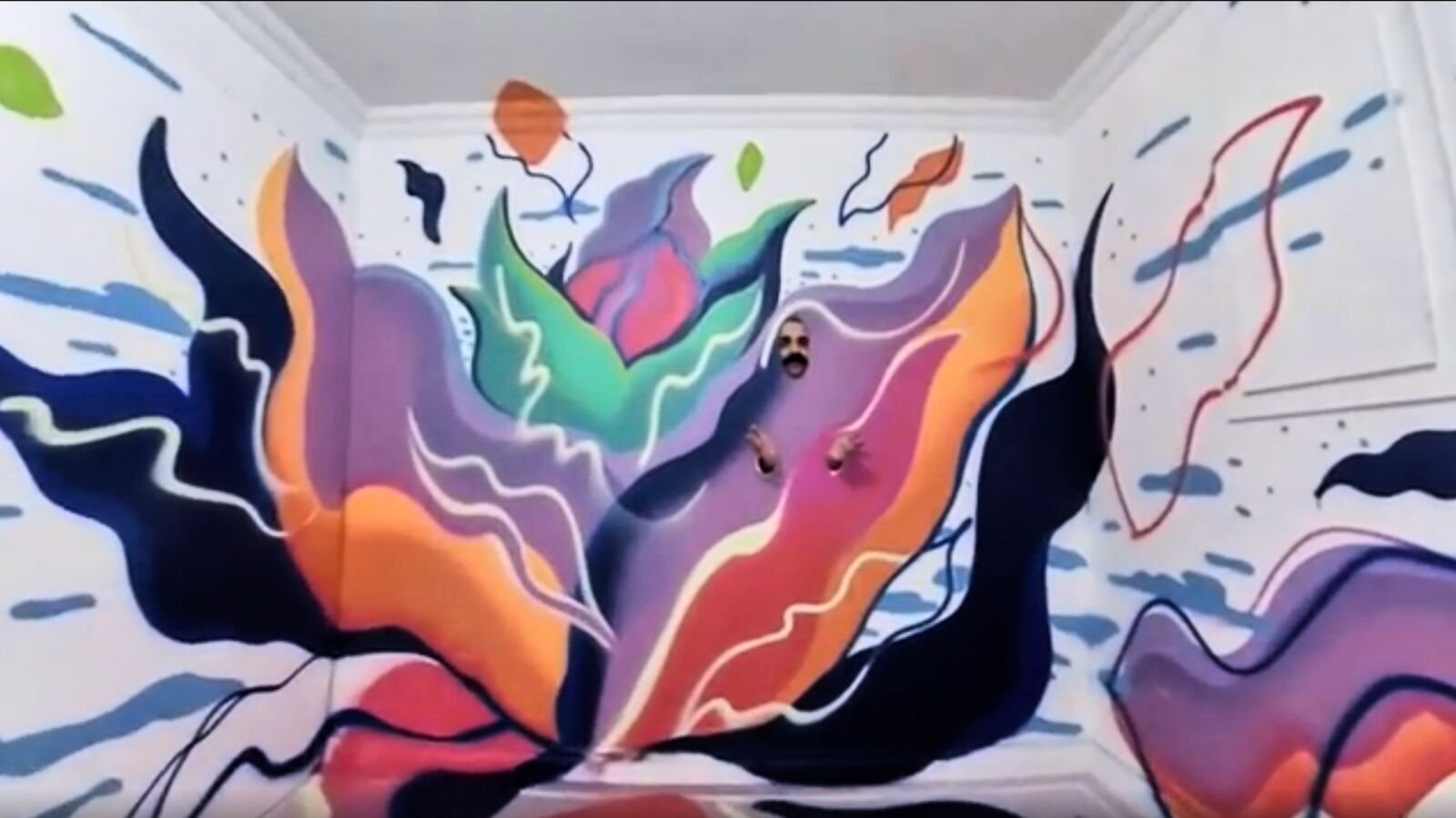 Raj Gill appearing within a colourful abstract mural painted across the walls of a room