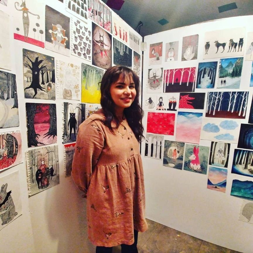 An artist standing in a corner of the gallery with walls covered in small illustrated paintings