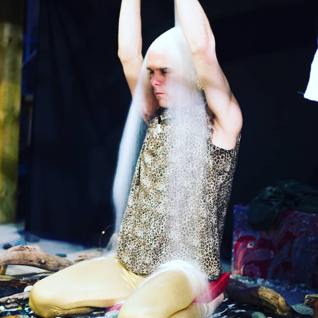 A performer seated cross-legged in a leopard-print shirt with motion-blurred arms raised overhead