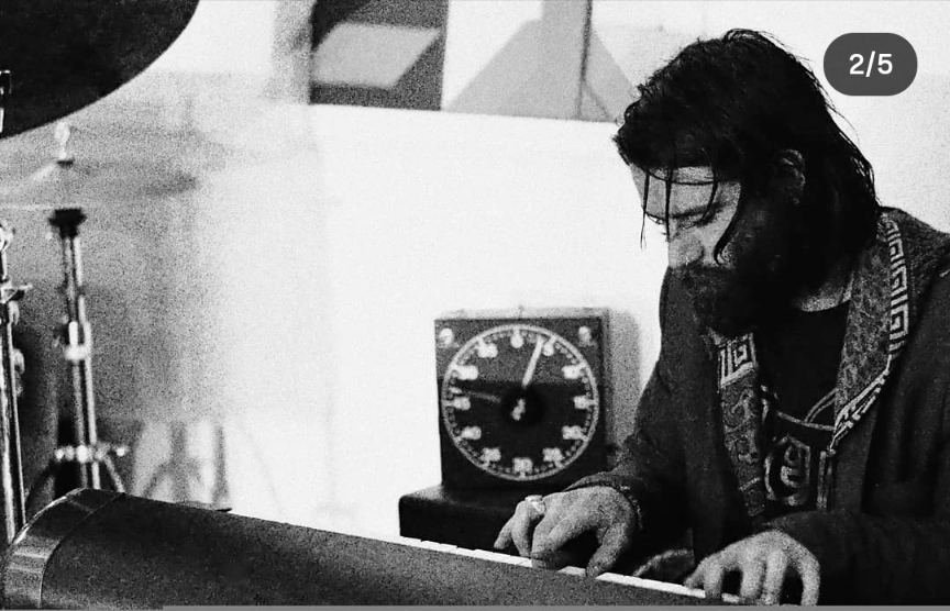 A musician playing keyboard, long hair falling forward, a clock on the wall behind